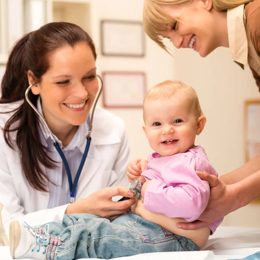 Foto einer Untersuchung beim Kinderarzt – fröhliches Baby auf der Untersuchungsliege, lächelnde Kinderärztin mit Stethoskop und liebevolle Mutter daneben