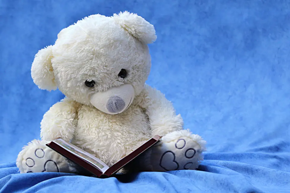 Foto eines kuscheligen, weißen Teddybären auf einer weichen blauen Decke, der vor einem geöffneten Buch sitzt – klassische Einschlafrituale wie Vorlesen